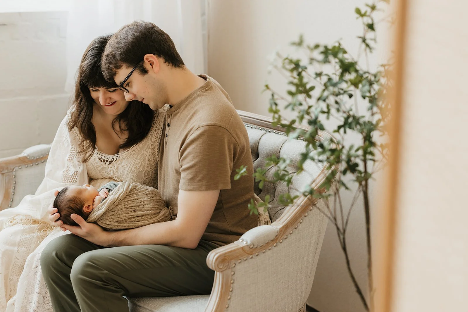 A couple sitting on a couch holding a newborn baby wrapped in a blanket, with a small plant in the foreground.