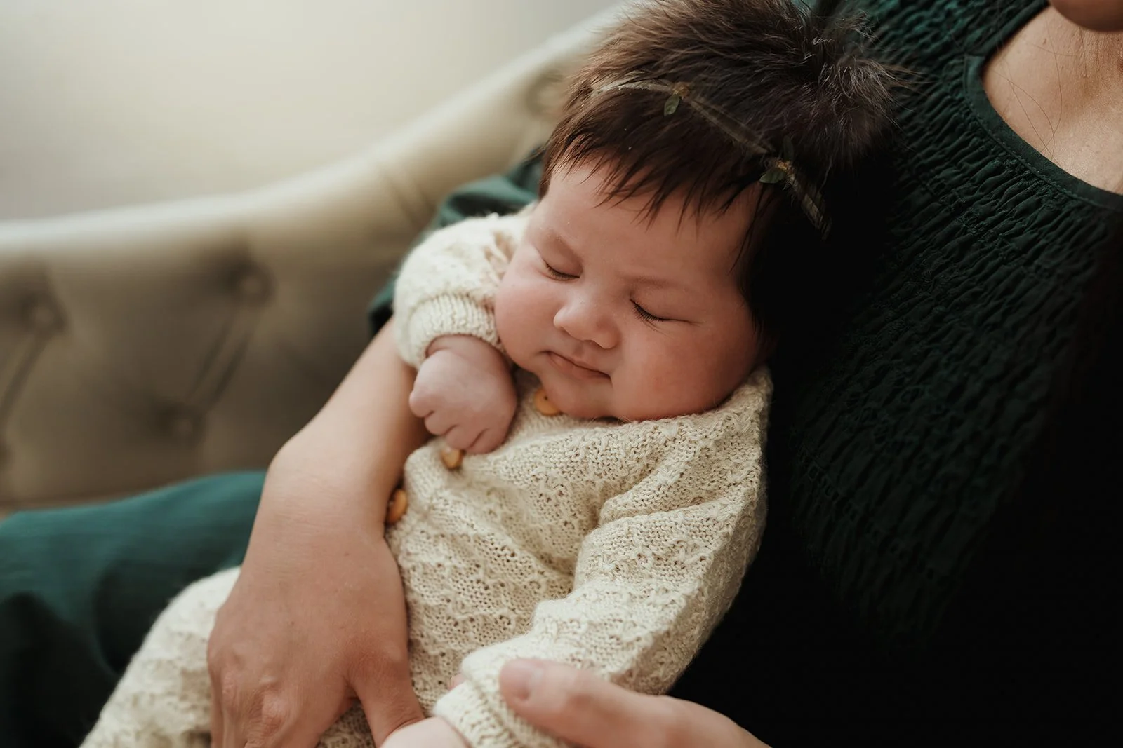 A sleeping baby resting on an adult's arm, wearing a cream-colored knit sweater, with their head supported on the adult's chest.