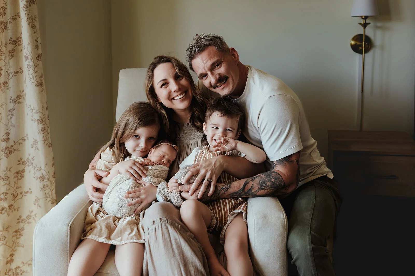 A happy multigeneration family sitting on a cream-colored armchair in a living room, smiling and cuddling together, including two young children, a newborn baby, a woman, and a man.
