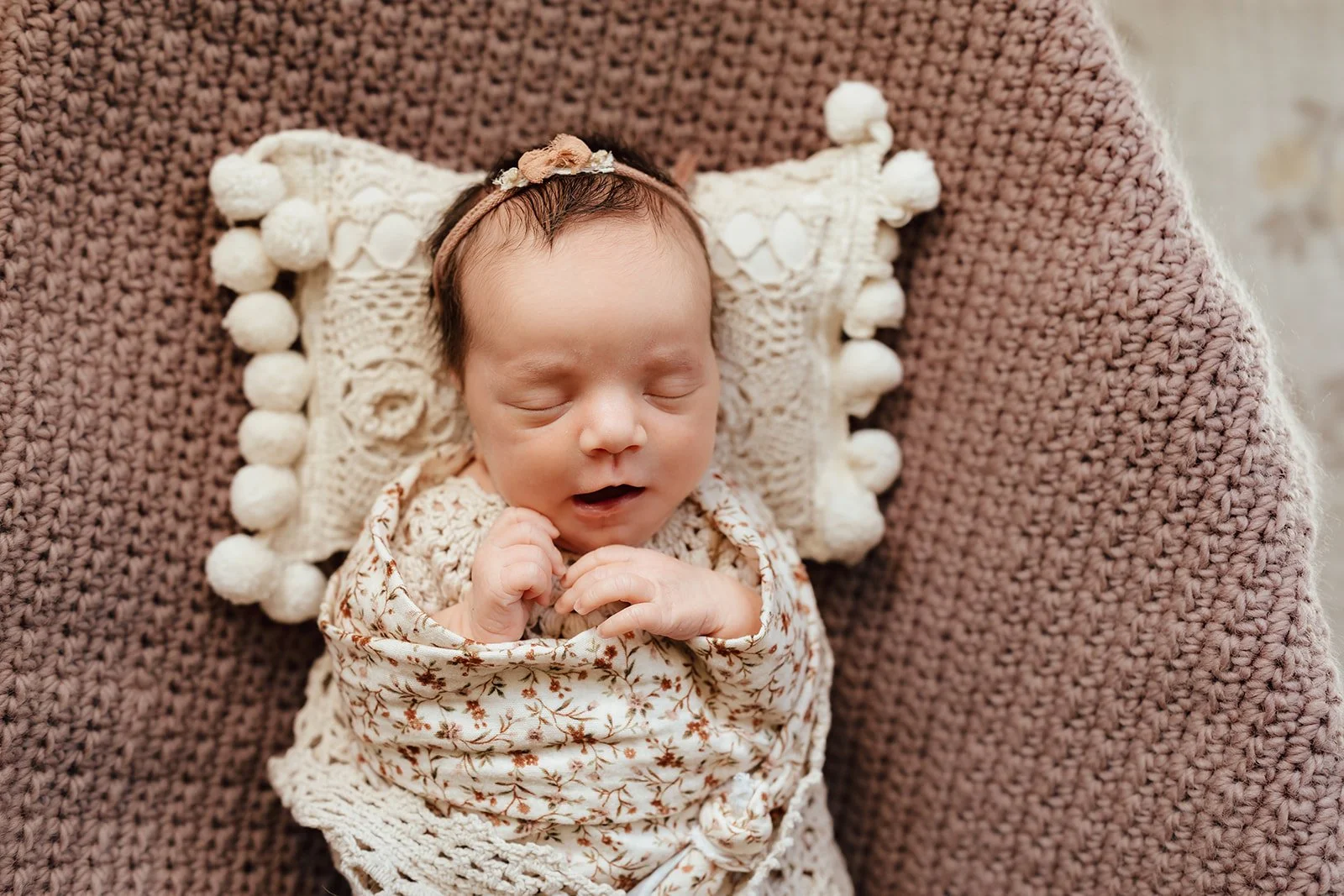 A sleeping newborn baby wrapped in a floral blanket, lying on a decorative crochet pillow with pom-poms, on a chunky knit blanket.
