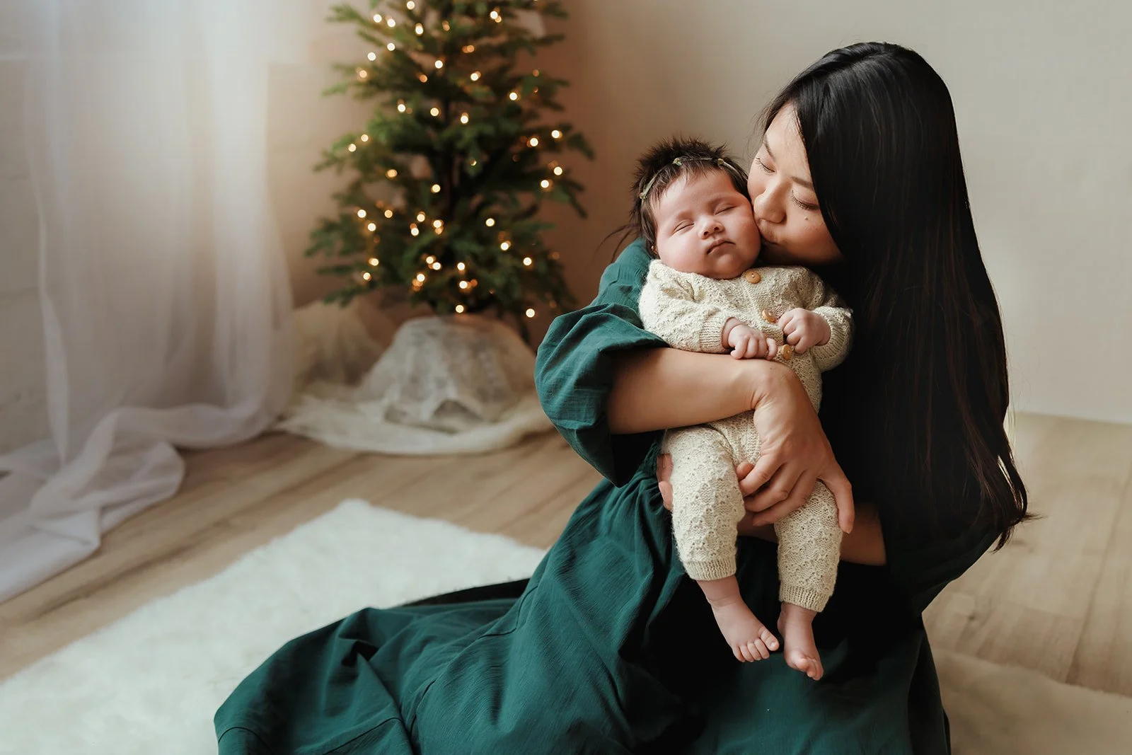 A woman with long dark hair is holding a sleeping baby dressed in cream-colored knit clothing, kissing the baby's head, with a decorated Christmas tree in the background.