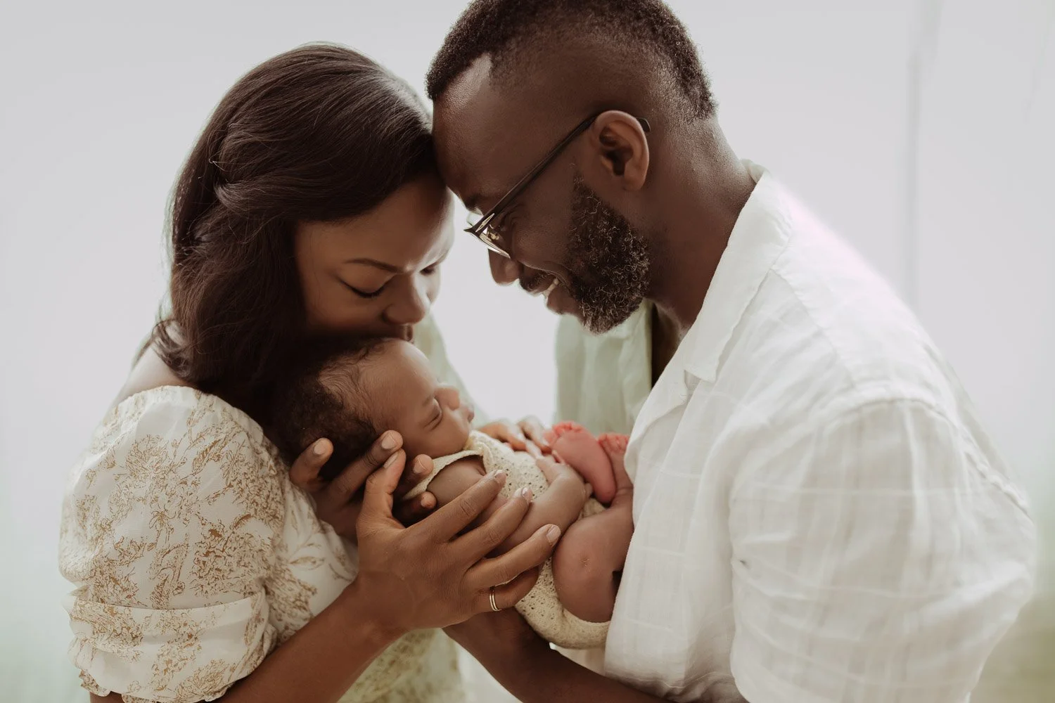 A mother and father stand in front of a floor to ceiling window with white curtains and hold their sleeping newborn baby girl between them.