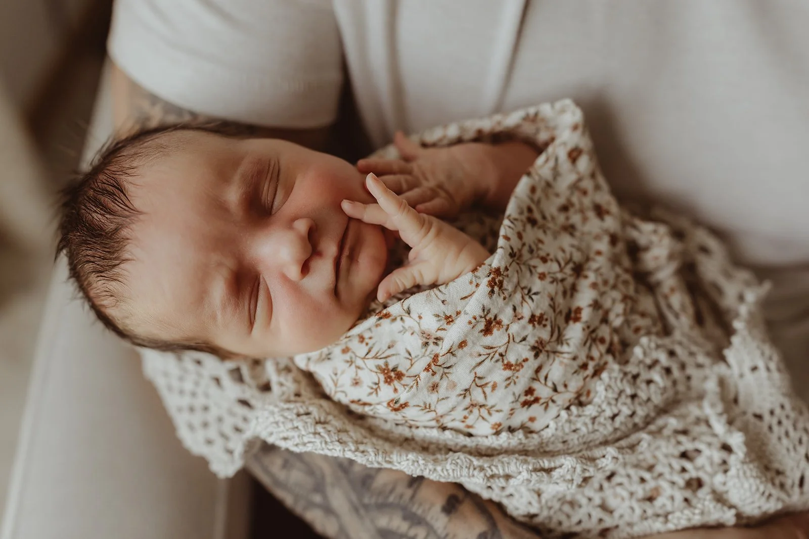 A newborn baby peacefully sleeping, wrapped in a floral-patterned blanket and lying on a crocheted cloth.