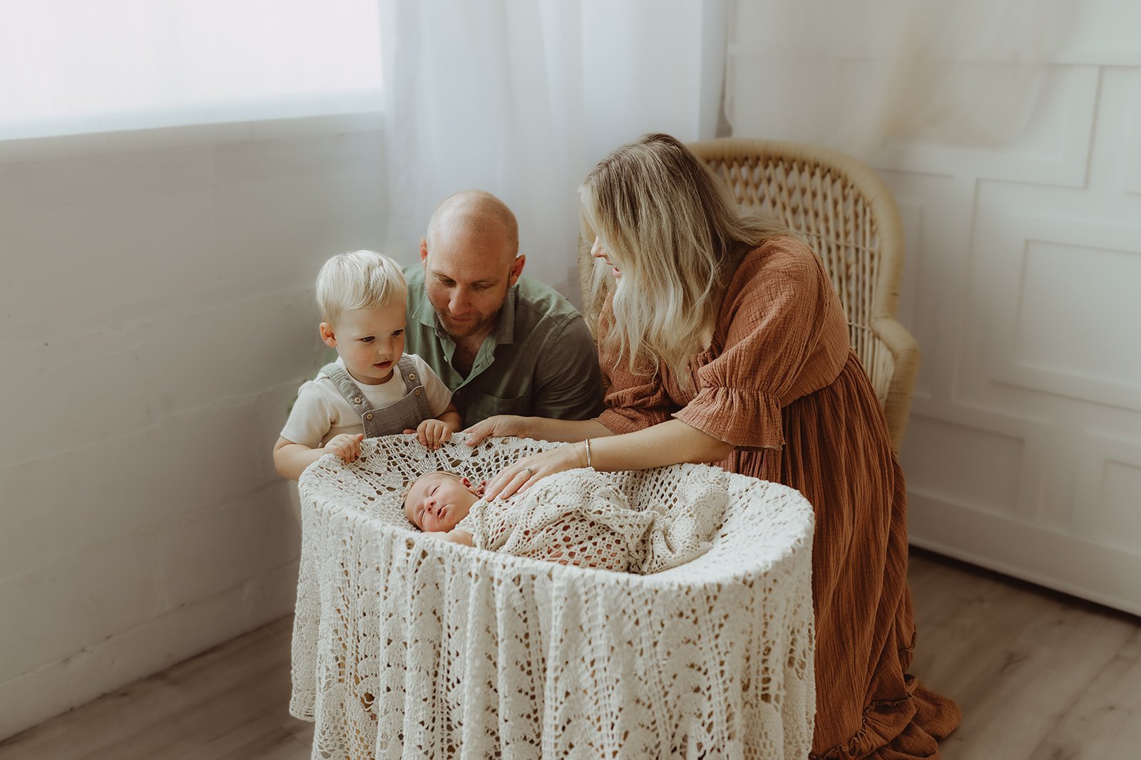 a dad and mom are showing their toddler son his new baby sister who is sleeping in a bassinet