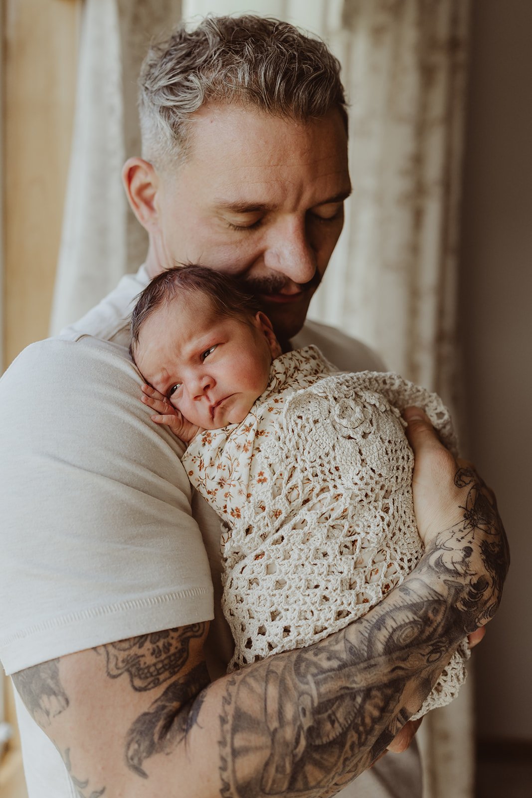A man with tattoos on his arms holding a newborn baby wrapped in a crocheted blanket, both indoors with curtains in the background.