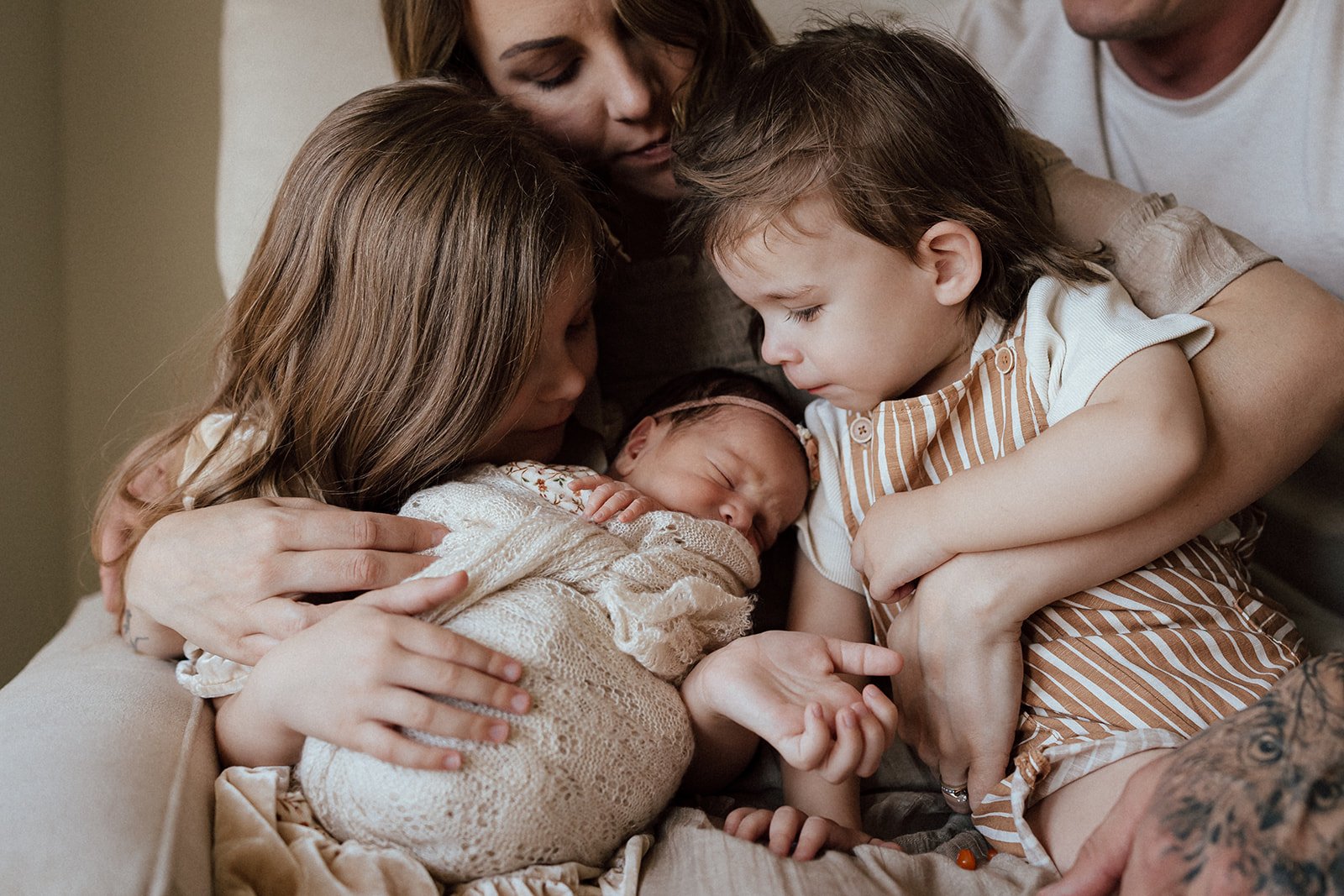 A family gathering around a newborn baby in a cozy setting, with two young children and two adults softly touching the baby's head and hand.