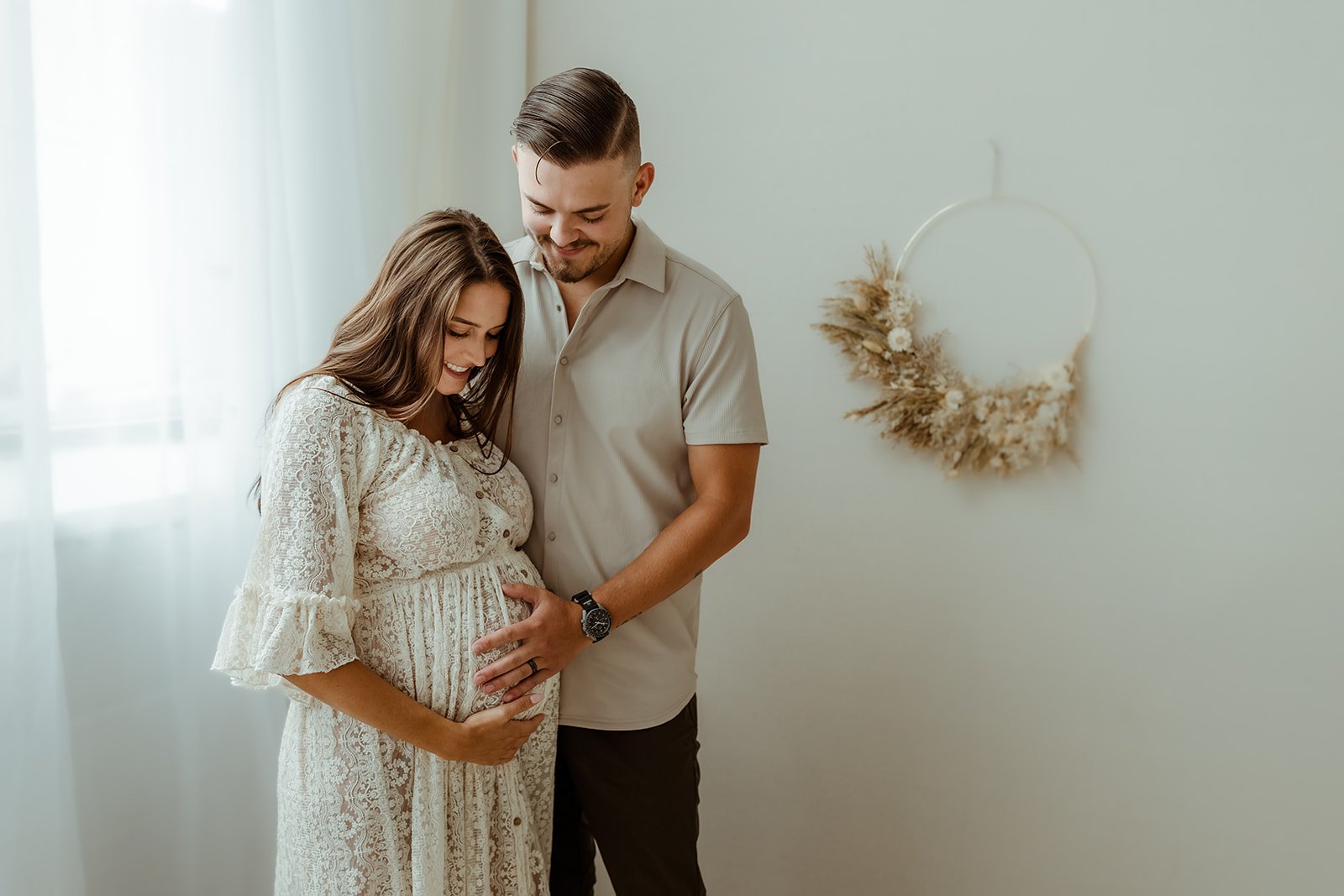 A pregnant woman and her partner standing in a bright room, smiling and gently holding her belly, with a minimalist wall hanging in the background.