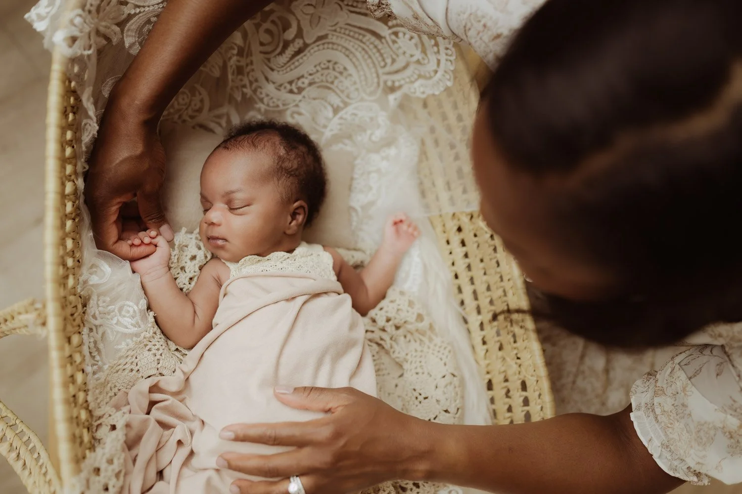 A mother leans over the bassinet where her baby girl sleeps and holds her hand during their newborn photography session.