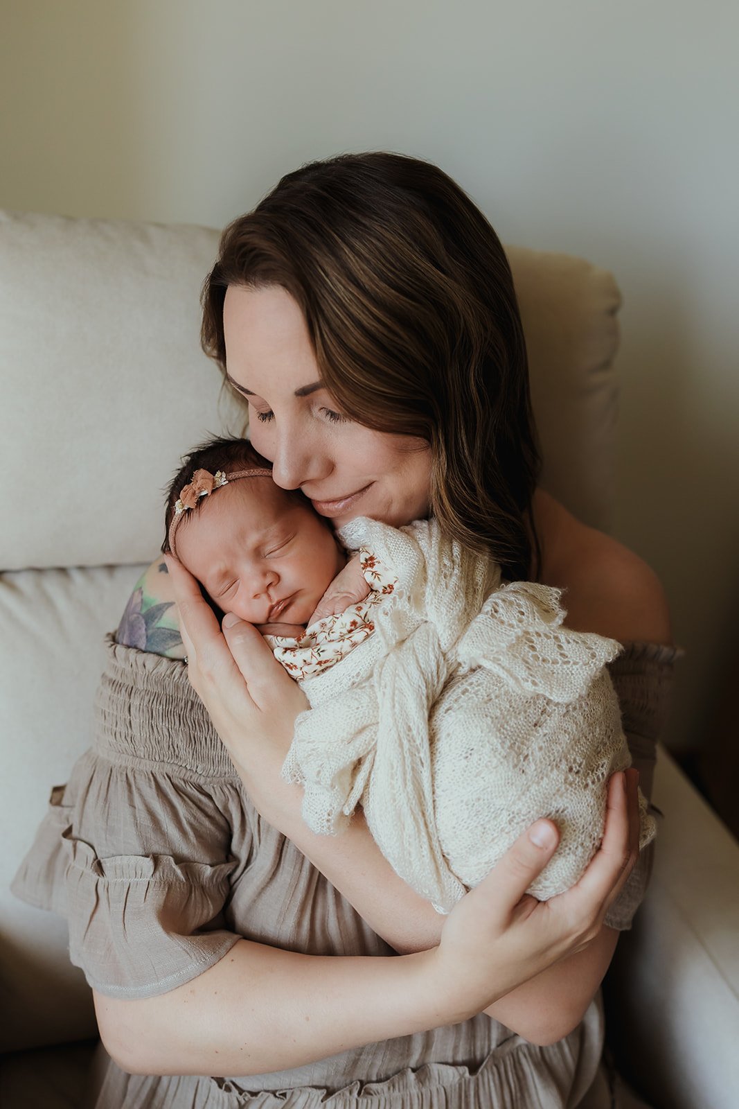 A woman gently holds a sleeping newborn baby close to her chest, with her eyes closed and a gentle smile. The baby is wrapped in a cream-colored blanket with lace and floral details and wears a small pink headband with flowers, resting peacefully.