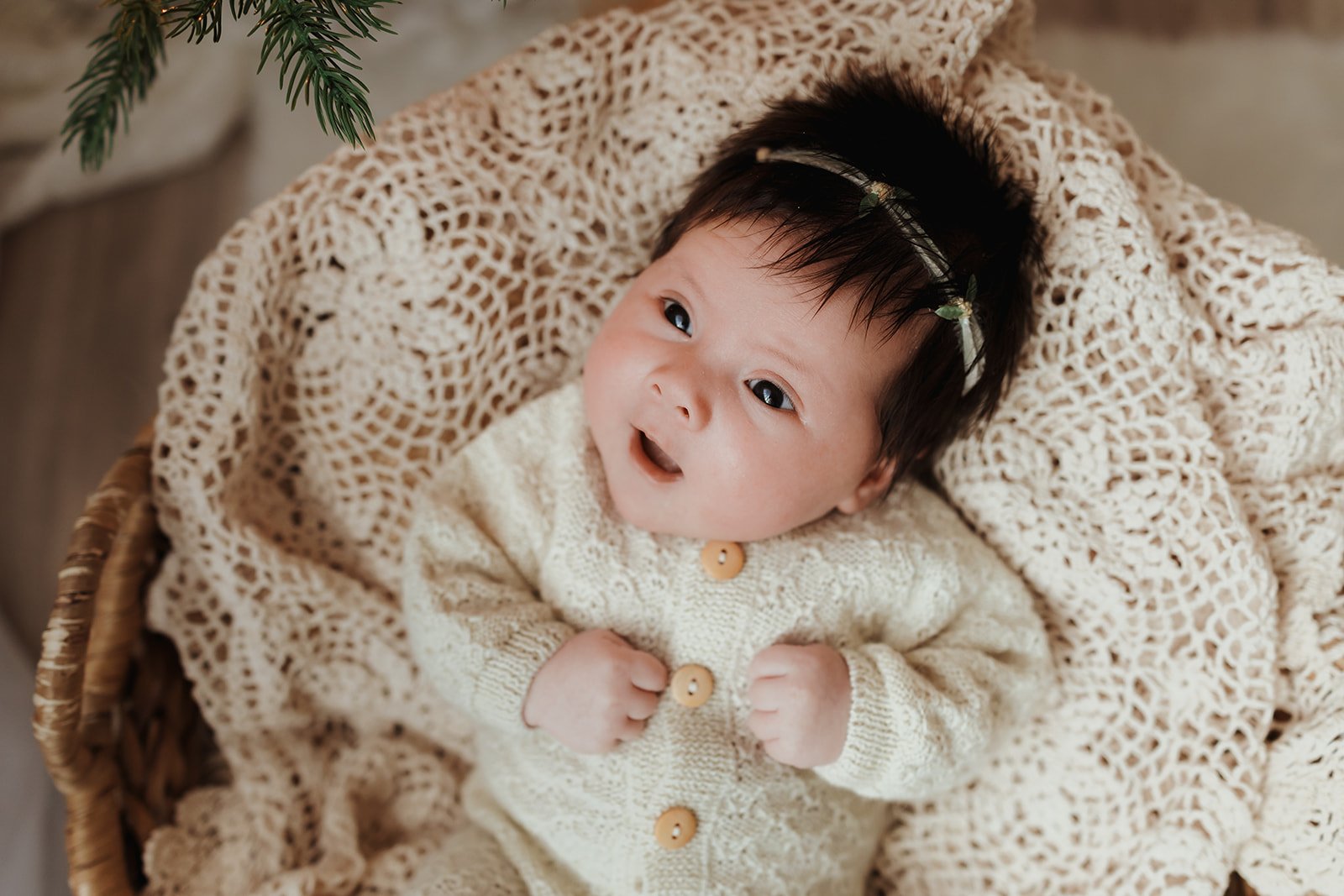 A baby with dark hair, lying on a knitted blanket in a wicker basket, looking up with a surprised expression, wearing a cream-colored knitted sweater with buttons, and a headband with small green leaves.
