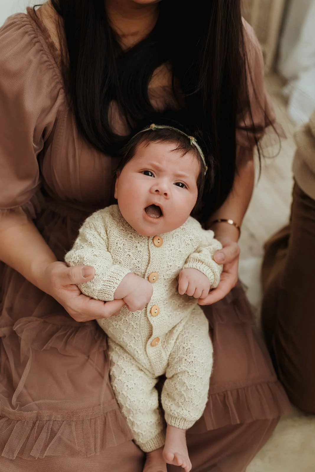 A woman holding a baby girl dressed in a cream-colored knitted outfit. The baby appears to be upset, with her mouth open and eyebrows furrowed.