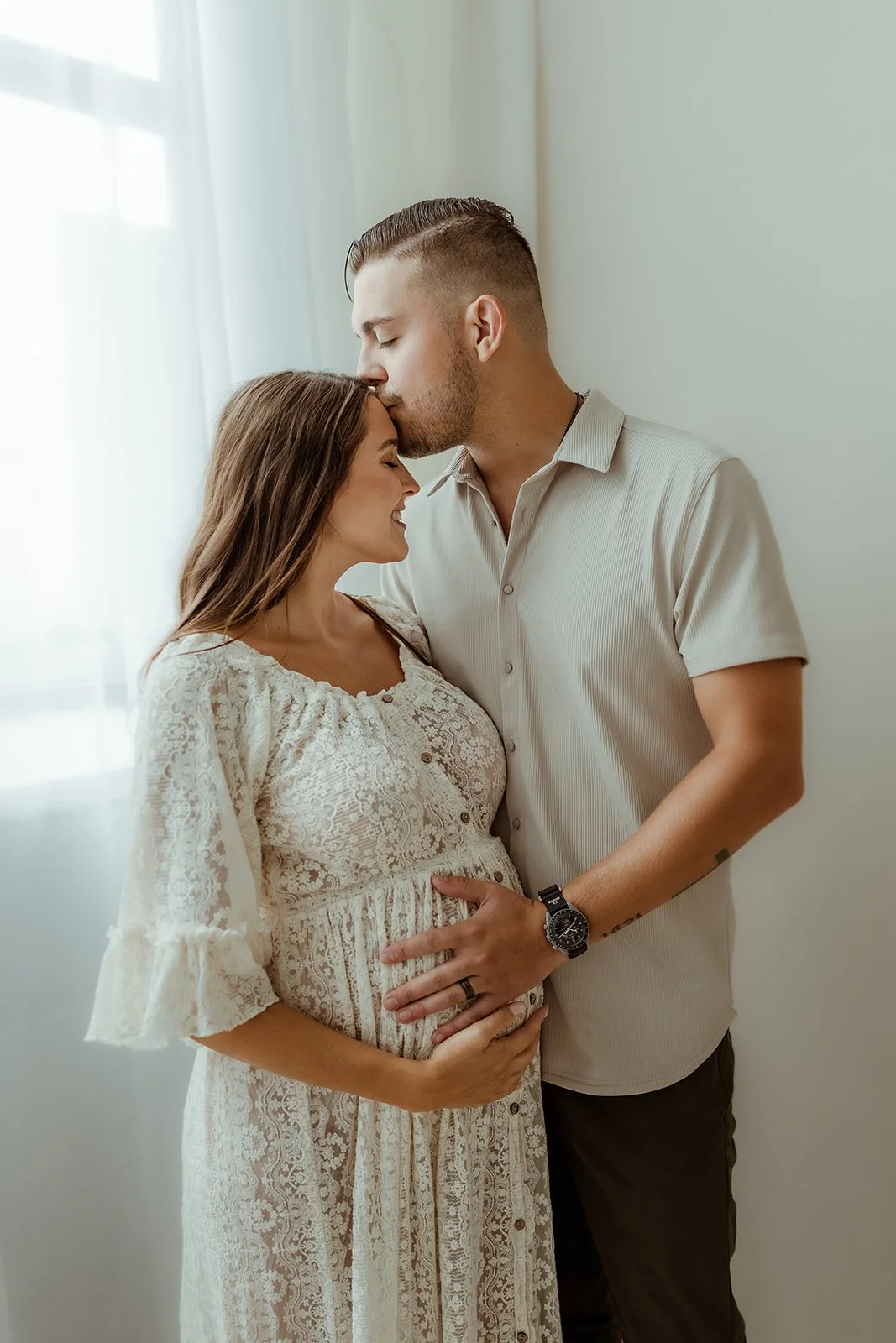 A pregnant woman and a man are standing close together indoors near a window. The man is kissing the woman's forehead while both are smiling. The woman is wearing a white lace dress, and the man is wearing a light-colored shirt with a watch on his wr