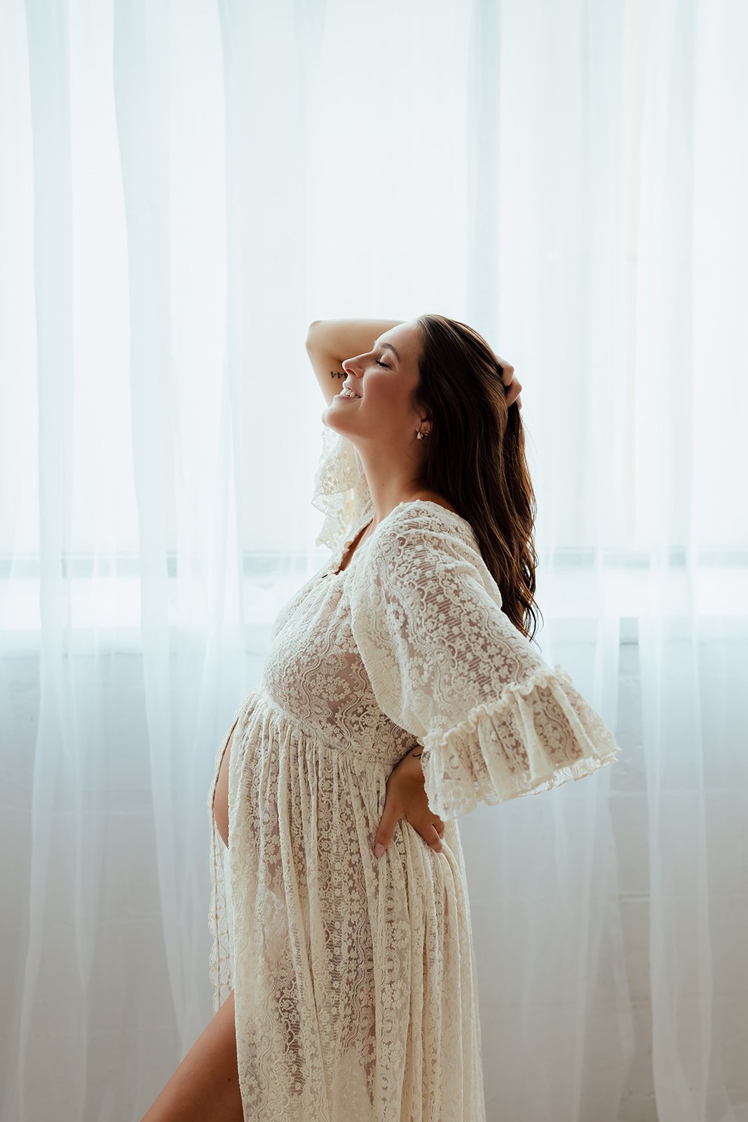 A pregnant woman smiling with her eyes closed, standing in front of white curtains, wearing a lace dress with her hand on her hip and the other arm resting on her head.
