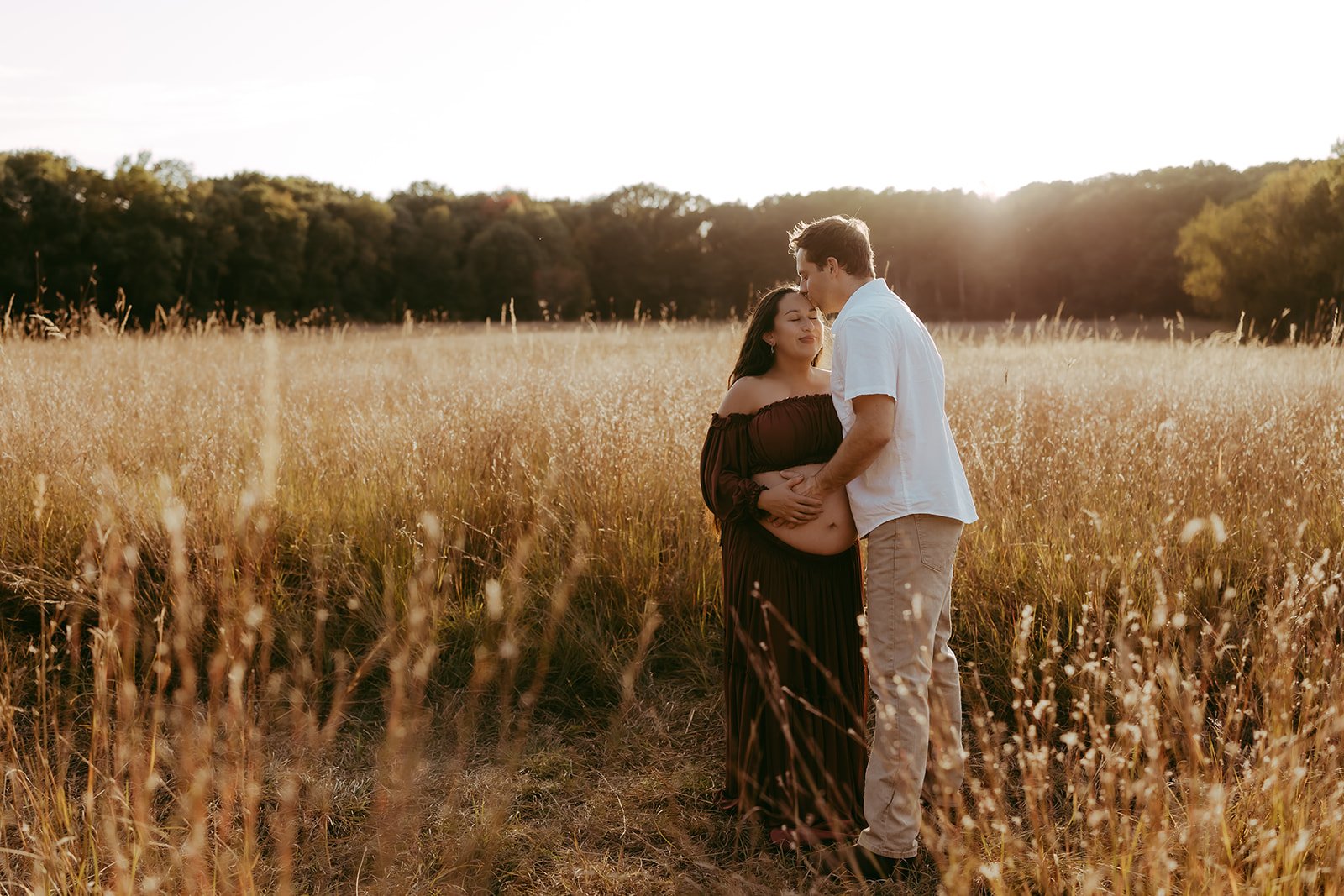 A couple stands in a field of tall, golden grass during sunset, with the man gently holding the woman's pregnant belly and touching her forehead, both with eyes closed.