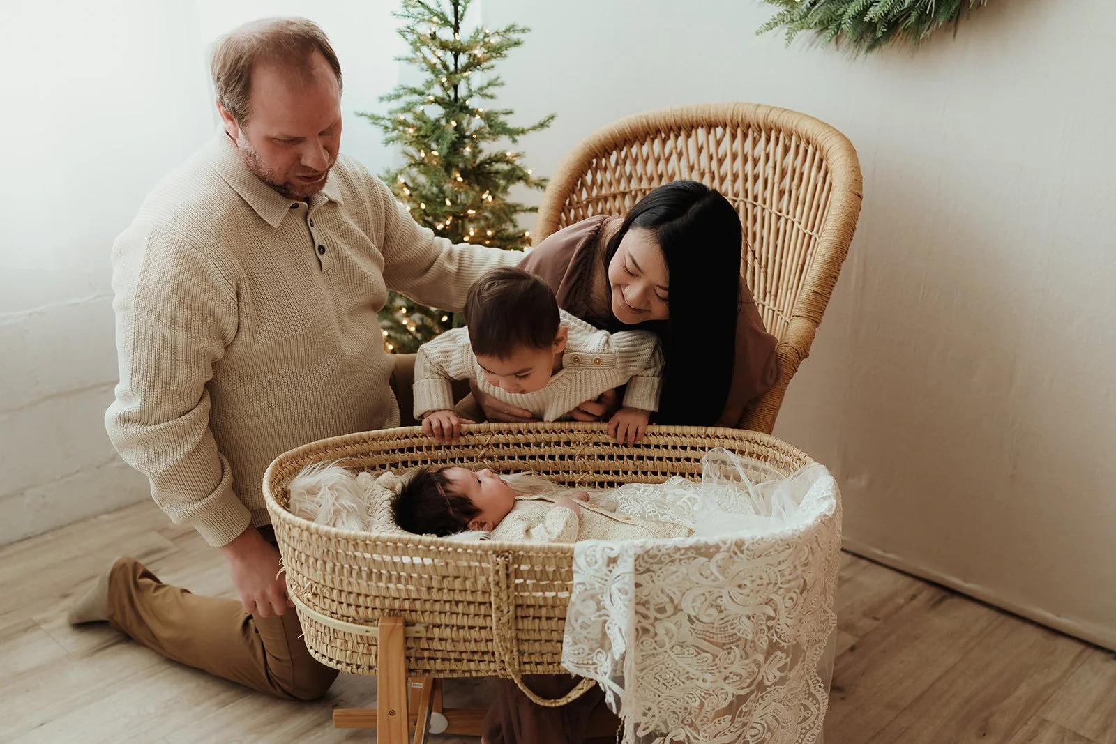 A family of four gathered around a baby in a bassinet during Christmas, with a decorated Christmas tree in the background.