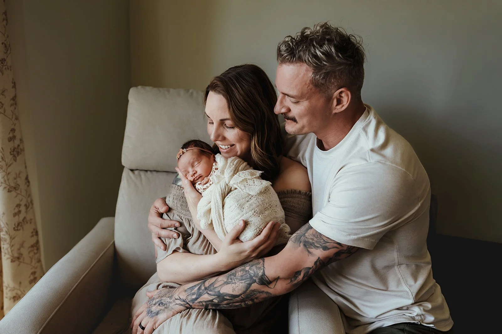 Parents holding their newborn baby on a beige couch in a cozy room, smiling and looking at the baby.
