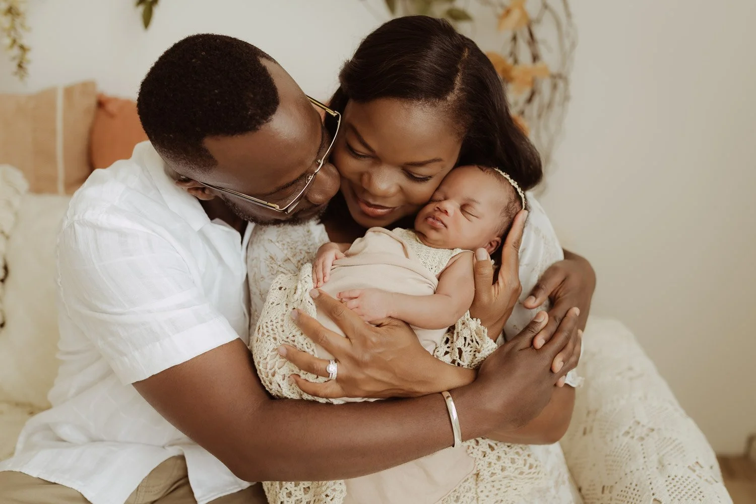 A mother and father sit together on the edge of a bed and hold their newborn baby girl between them.