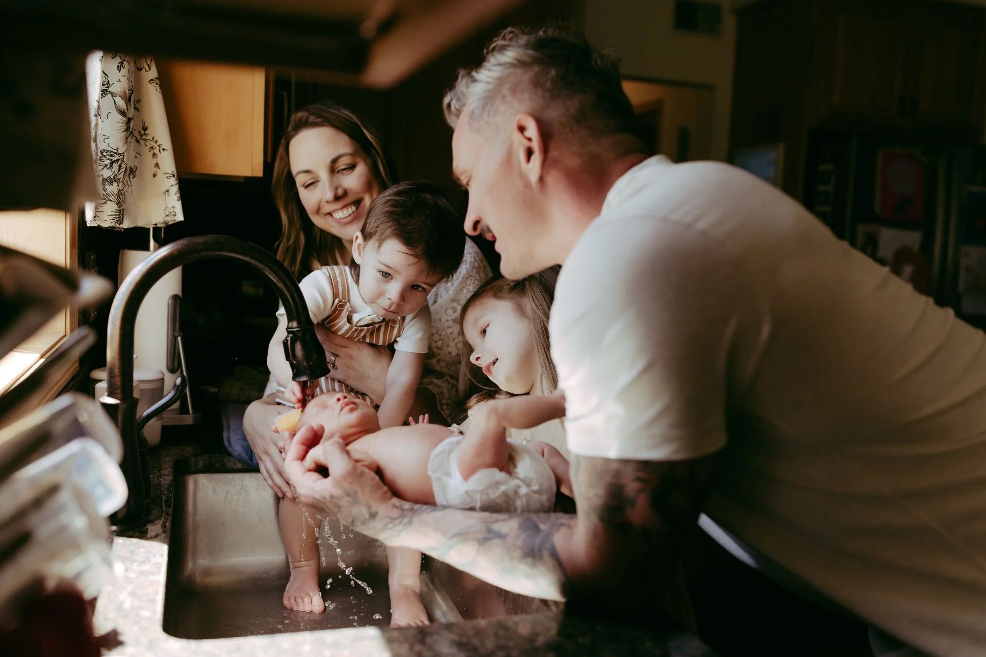 a family of four is washing their newborn baby's hair in their kitchen sink during a newborn photo shoot with AMG Photography