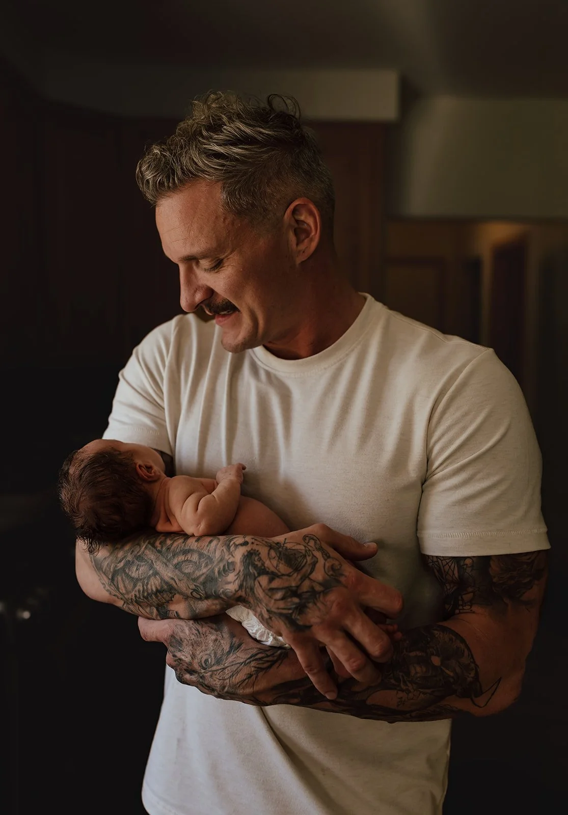 A man with tattoos on his arms holding a newborn baby in a dimly lit room.