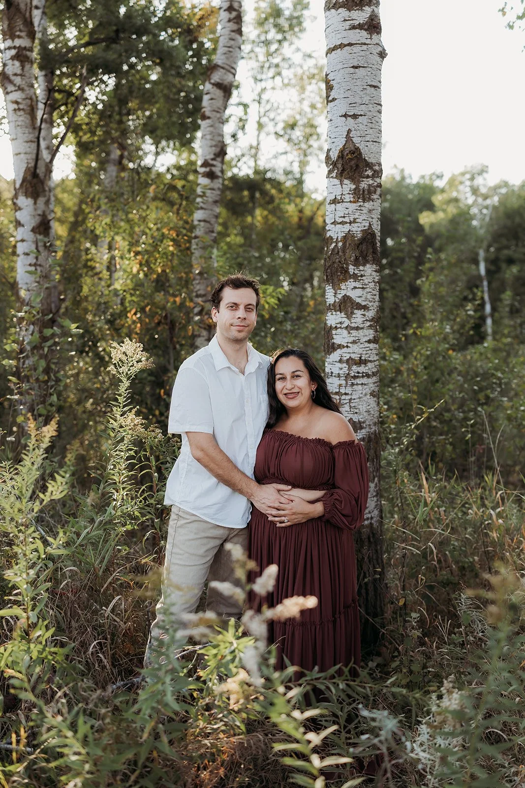 A pregnant woman and her partner standing together outdoors among trees and plants, with the woman wearing a long, off-the-shoulder brown dress and the man wearing a white shirt and beige pants, both smiling gently.