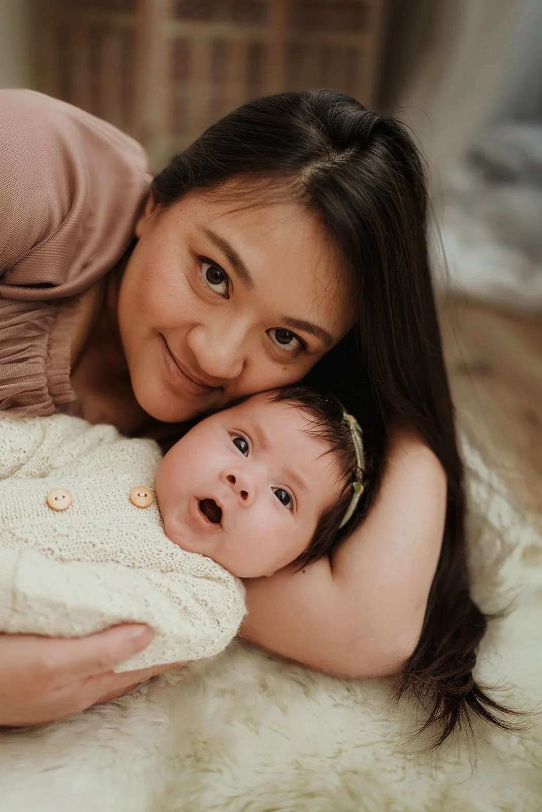 A woman with dark hair lying next to a baby with a bow headband, both smiling and looking at the camera, on a soft furry surface.