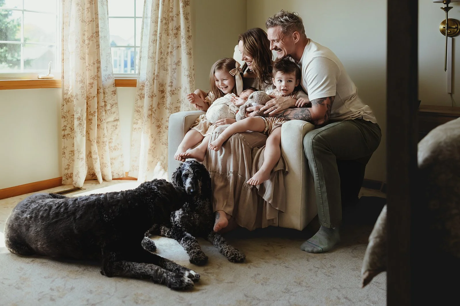 A happy family with three children and two dogs sitting on a beige sofa in a sunlit living room, laughing and playing together.