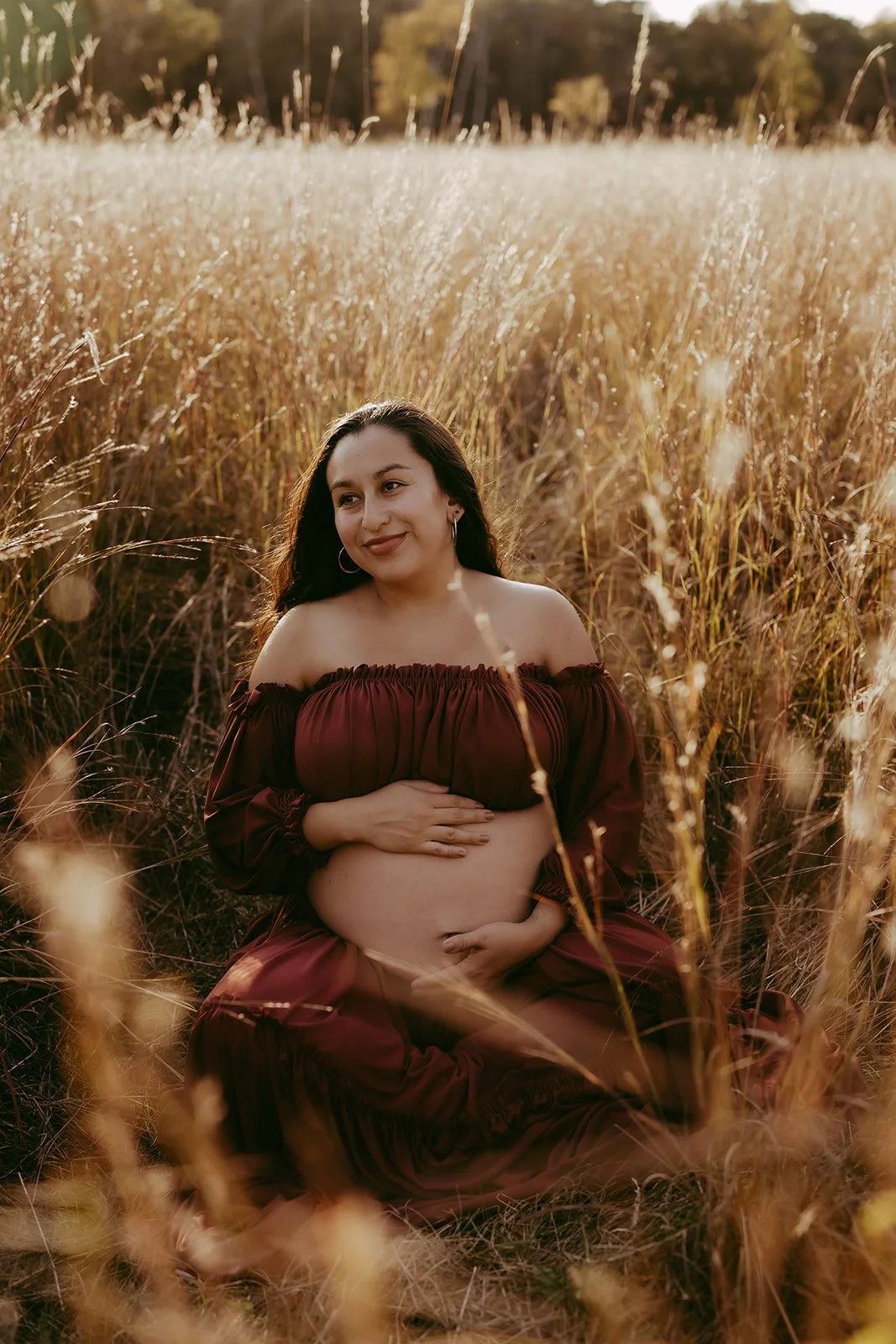 Pregnant woman sitting in a field of tall, golden grass during sunset, smiling softly and touching her pregnant belly.
