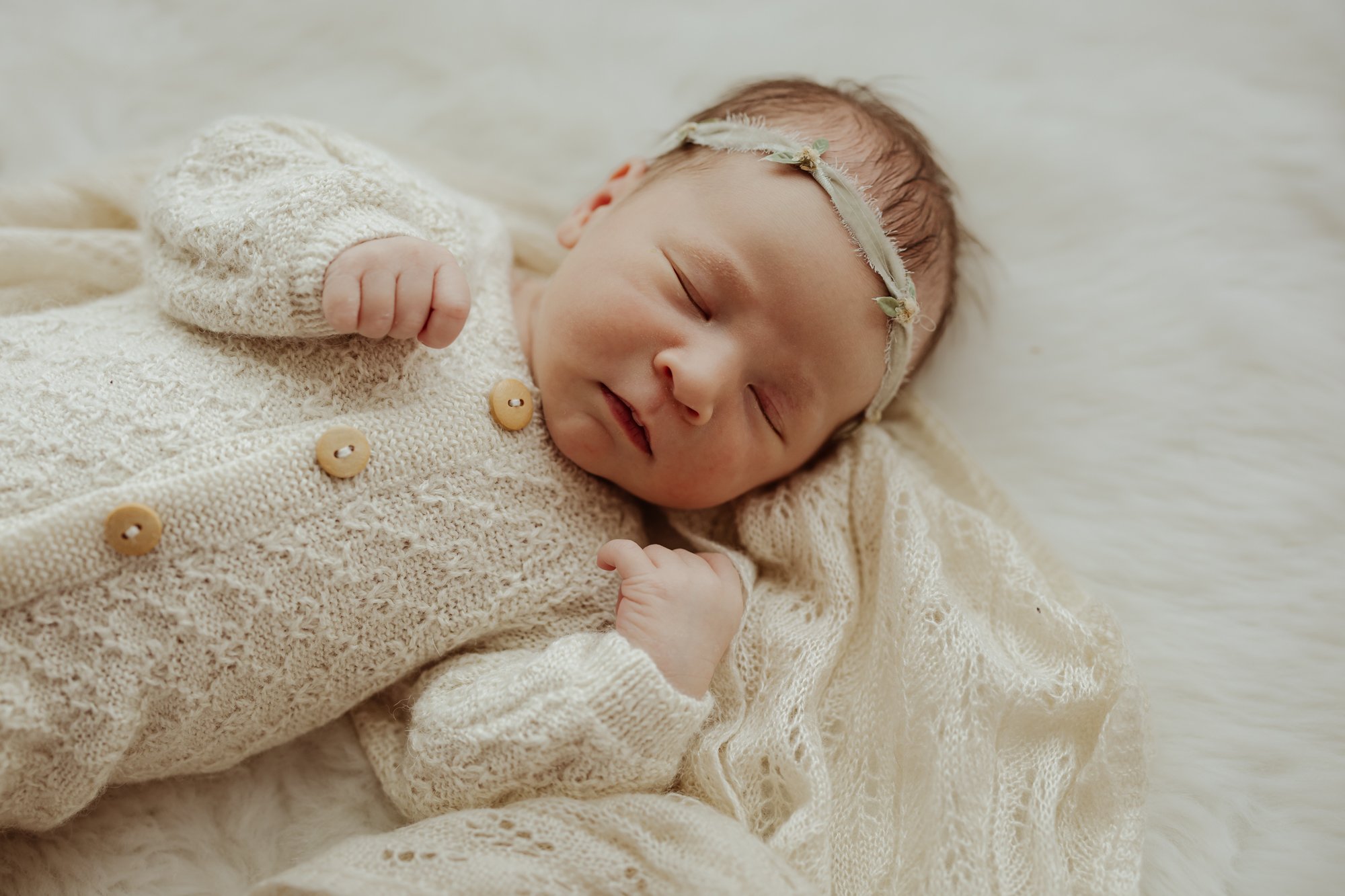 a baby girl wearing a cream colored hand knit jumper is sleeping on a soft cream colored blanket during her newborn photo shoot with AMG Photograqphy