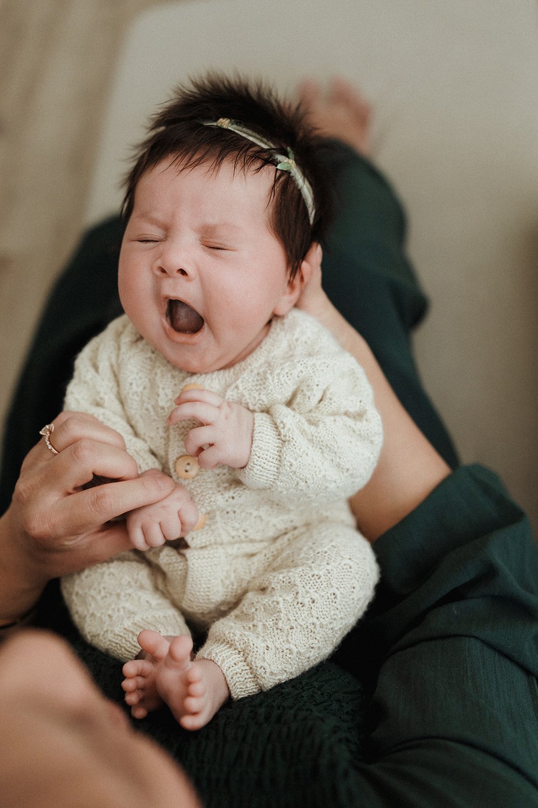 A yawning baby dressed in a cream knit onesie, lying on an adult's lap, in a cozy indoor setting.