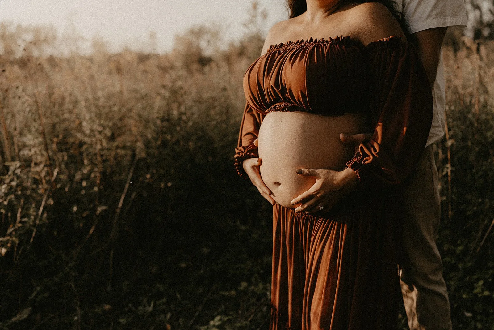 A pregnant woman standing outdoors with a man behind her, hands on her belly, during sunset in a field with tall grass.