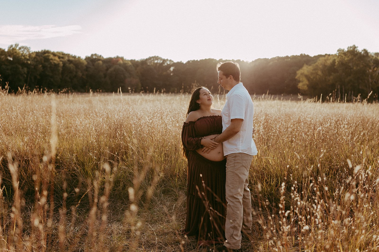 A pregnant woman and a man standing close together in a field of tall grass during sunset. The woman is wearing a dark off-the-shoulder dress and looking at the man, who is wearing a white shirt and beige pants. The sun is setting behind them, creati