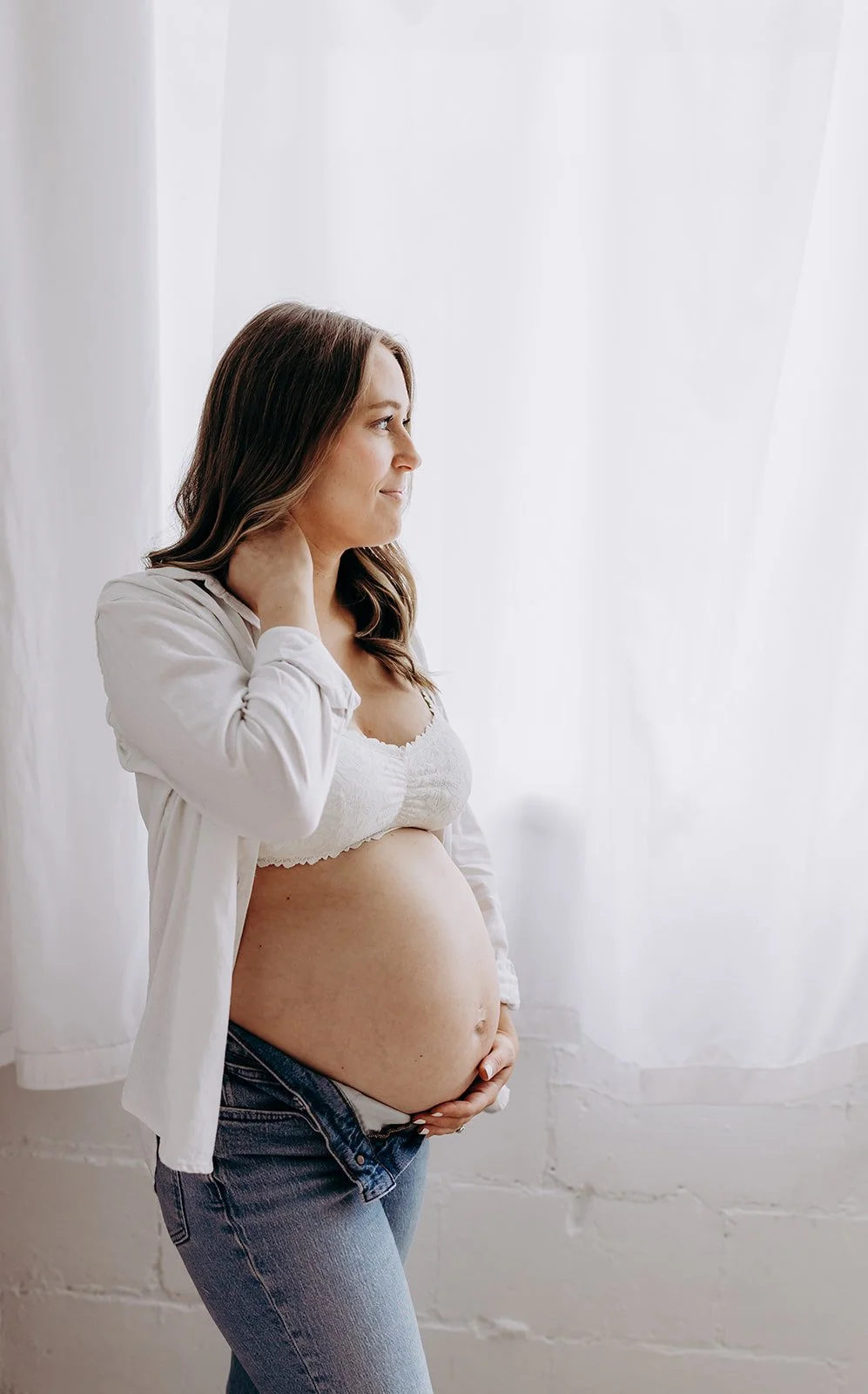 a pregnant woman in a white bralette and jeans is looking out a window at AMG Photography's studio during a maternity photo shoos.