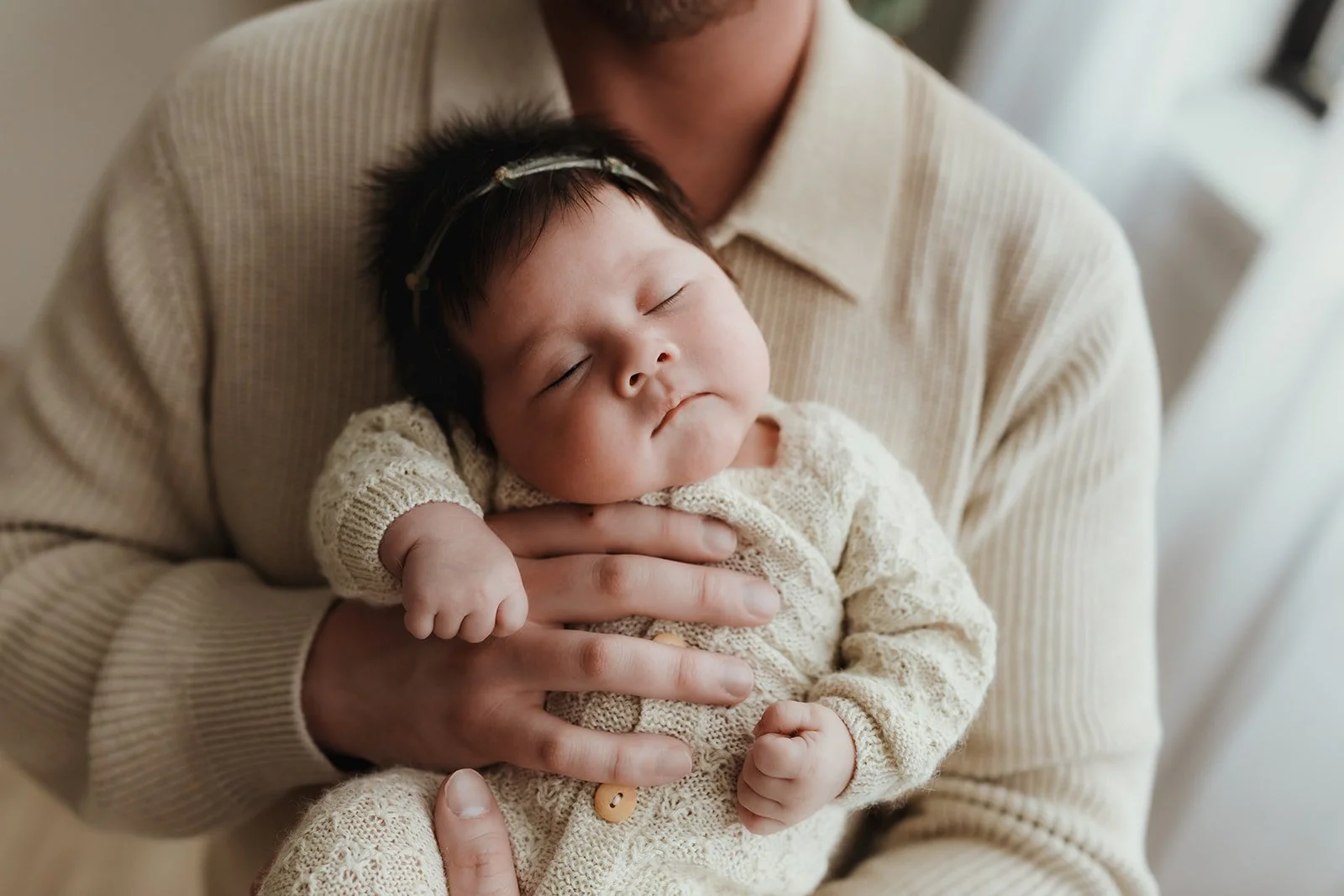 A sleeping baby with dark hair wearing a cream-colored knit outfit, being held by an adult person wearing a beige sweater.