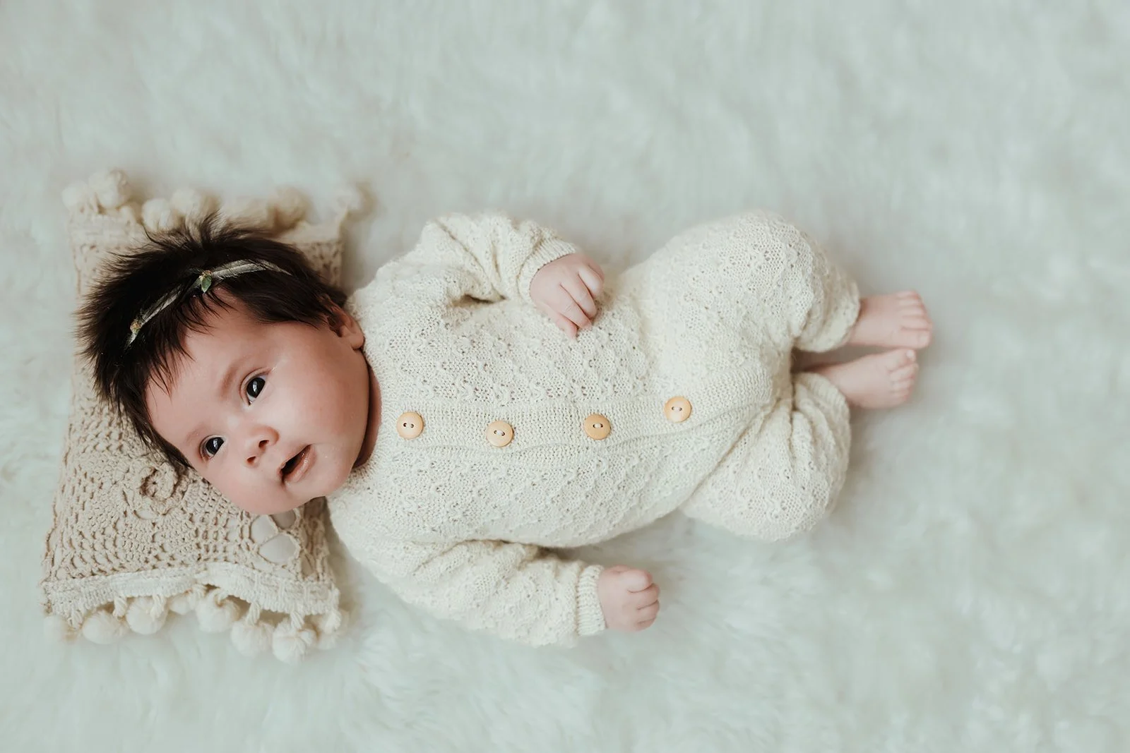 A baby lying on a soft white surface, wearing a cream-colored knitted outfit with buttons, resting on a crocheted pillow.