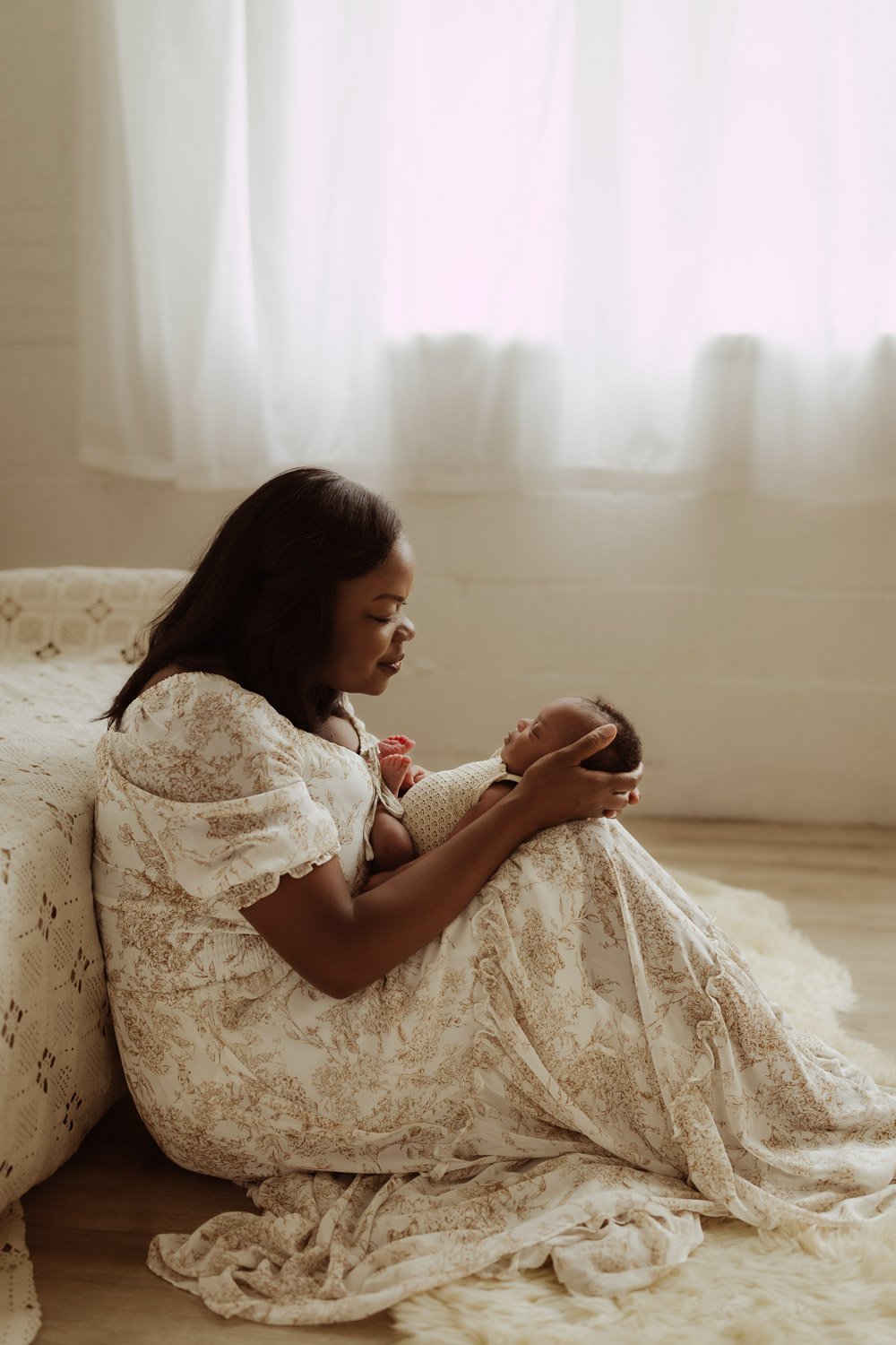 A mother sits at the foot of a bed covered by a lace blanket and holds her baby daughter in her arms.