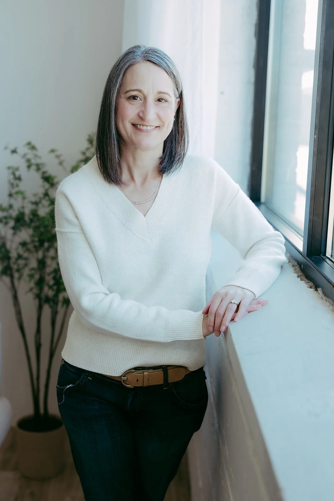 Mid-adult woman with shoulder-length dark hair smiling, leaning on a windowsill in a cozy indoor setting with a potted plant in the background.