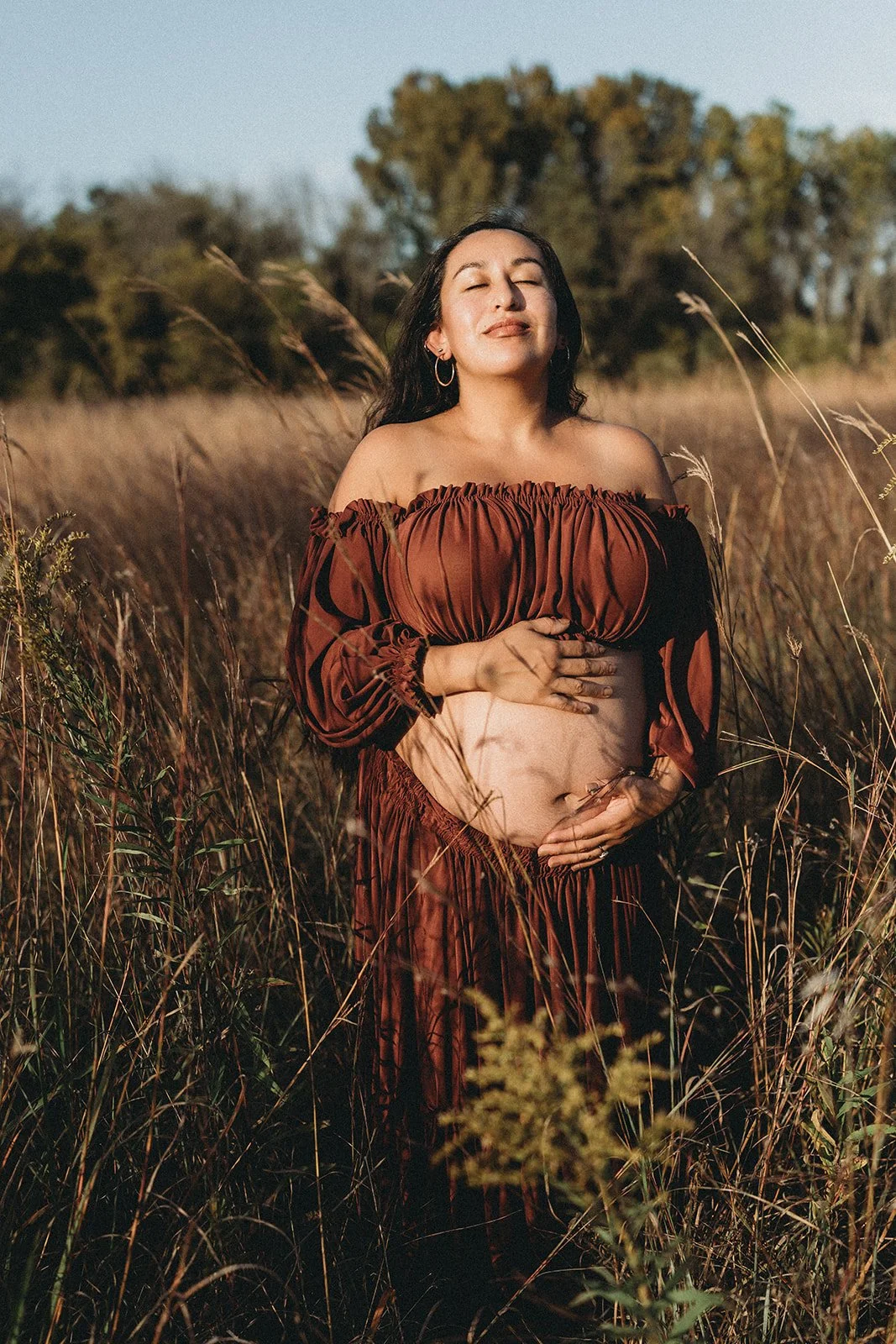 A pregnant woman in a brown off-shoulder dress standing in a field of tall grass with her eyes closed and hands on her belly.