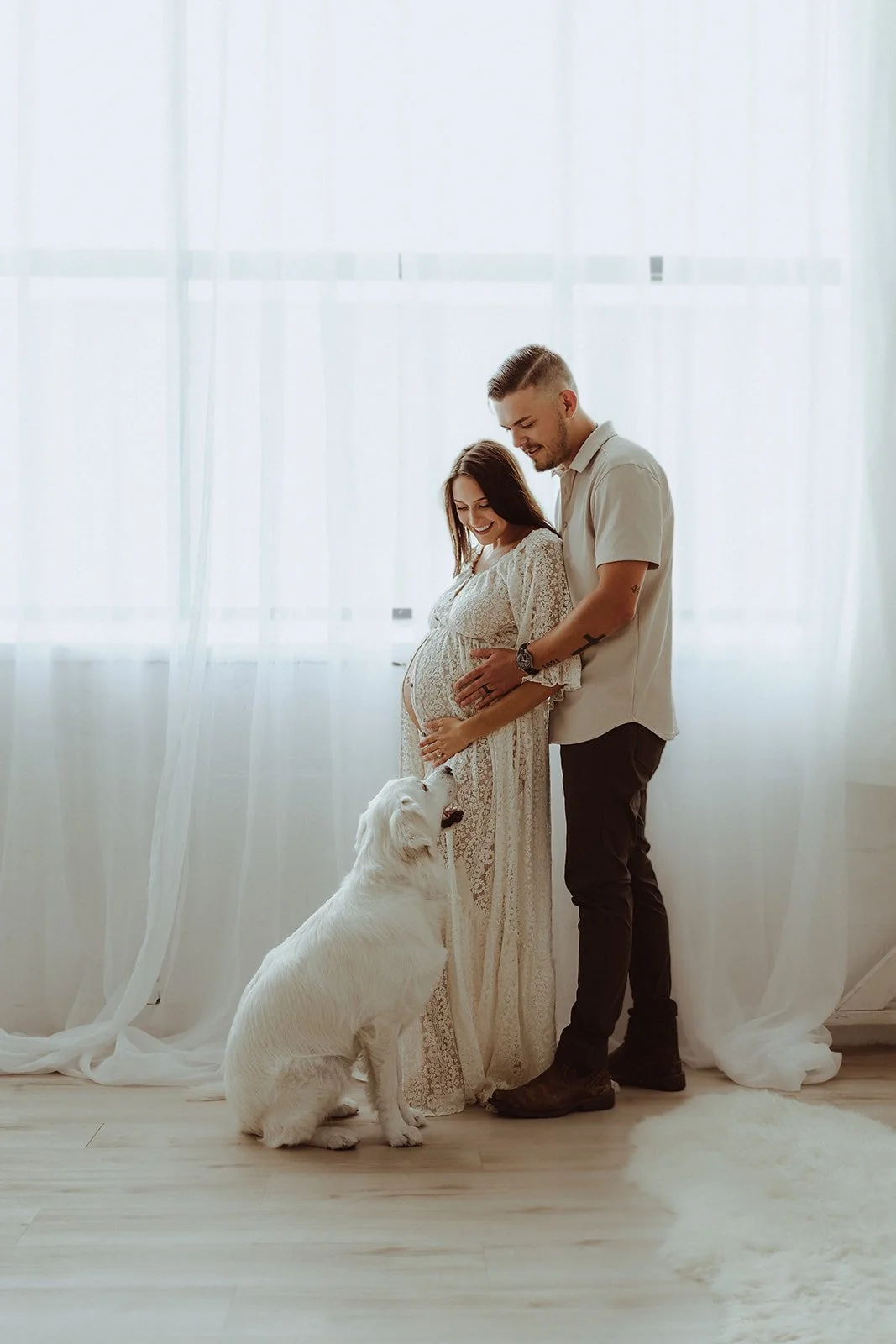 A pregnant woman in a lace dress touching her belly, smiling while a man standing beside her, touching her belly, smiling, and a white dog sitting on the floor, licking her hand, in front of a sheer curtain and wooden floor.