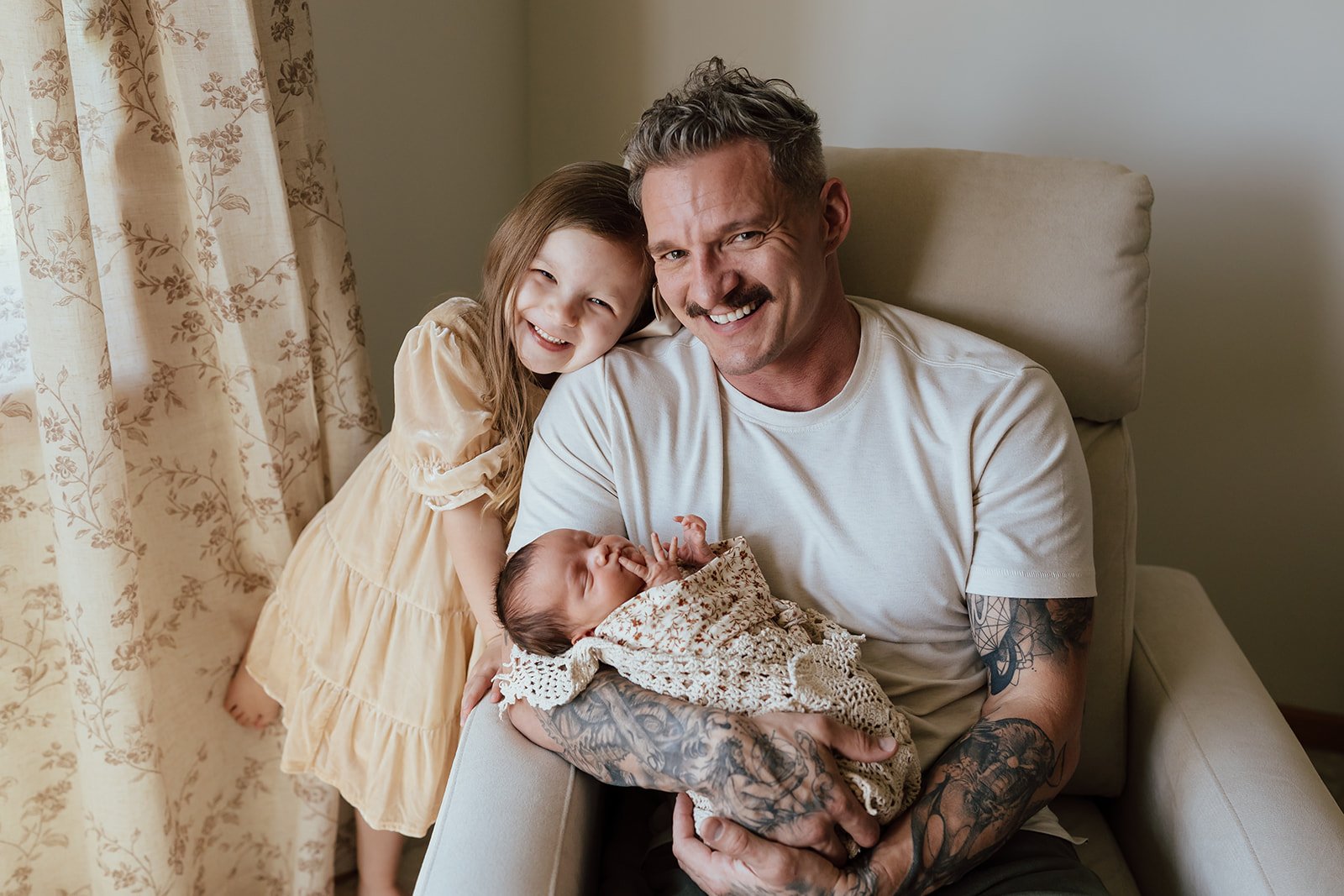 A smiling man with tattoos holding a newborn wrapped in a blanket, with a young girl leaning on his shoulder near floral curtains.