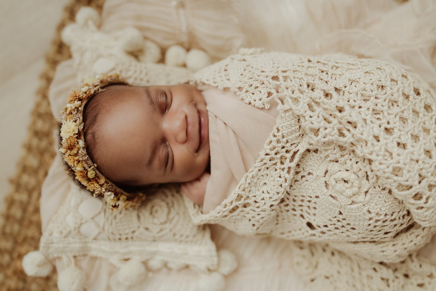 A newborn baby girl wearing a floral headband smiles in her sleep as she is wrapped in lacy blankets.