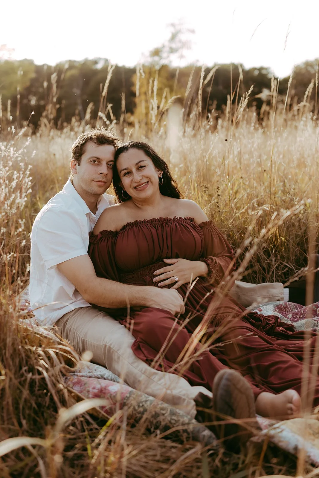 A couple sitting in a field of tall grass during sunset, with the woman pregnant and holding her belly, both smiling at the camera.