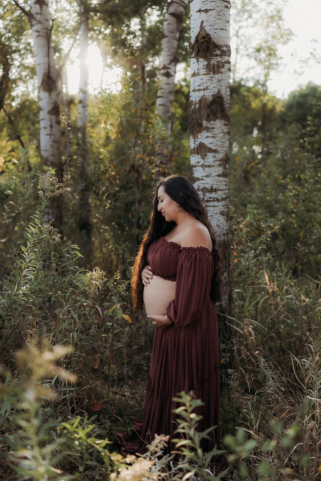 A pregnant woman in a burgundy maternity dress stands in a forest, touching her belly and looking down with a serene expression, sunlight filtering through trees.