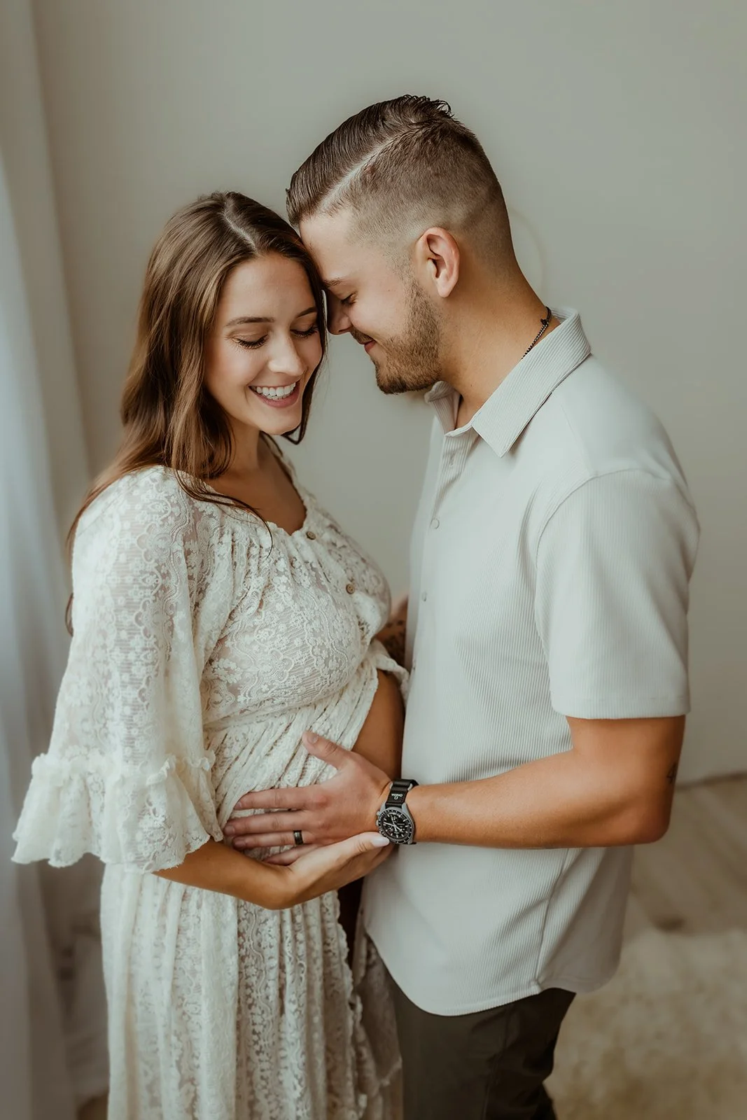 A happy couple with the woman pregnant, touching her belly, standing close together indoors.