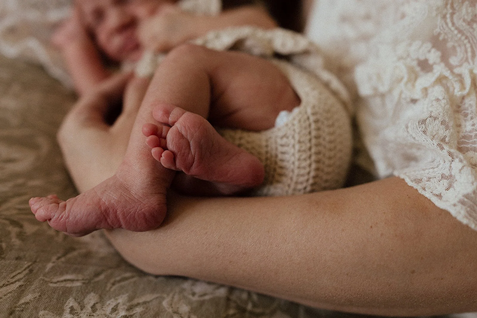 A newborn baby holding an adult's finger while lying on a lace-covered surface, wearing a knitted outfit.
