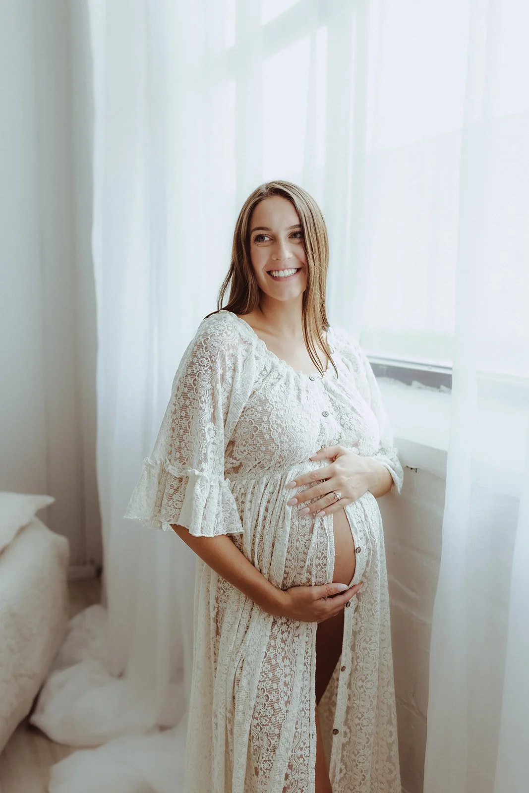 A pregnant woman with light brown hair wearing a white lace dress, standing near a window with sheer white curtains, smiling and gently touching her belly.