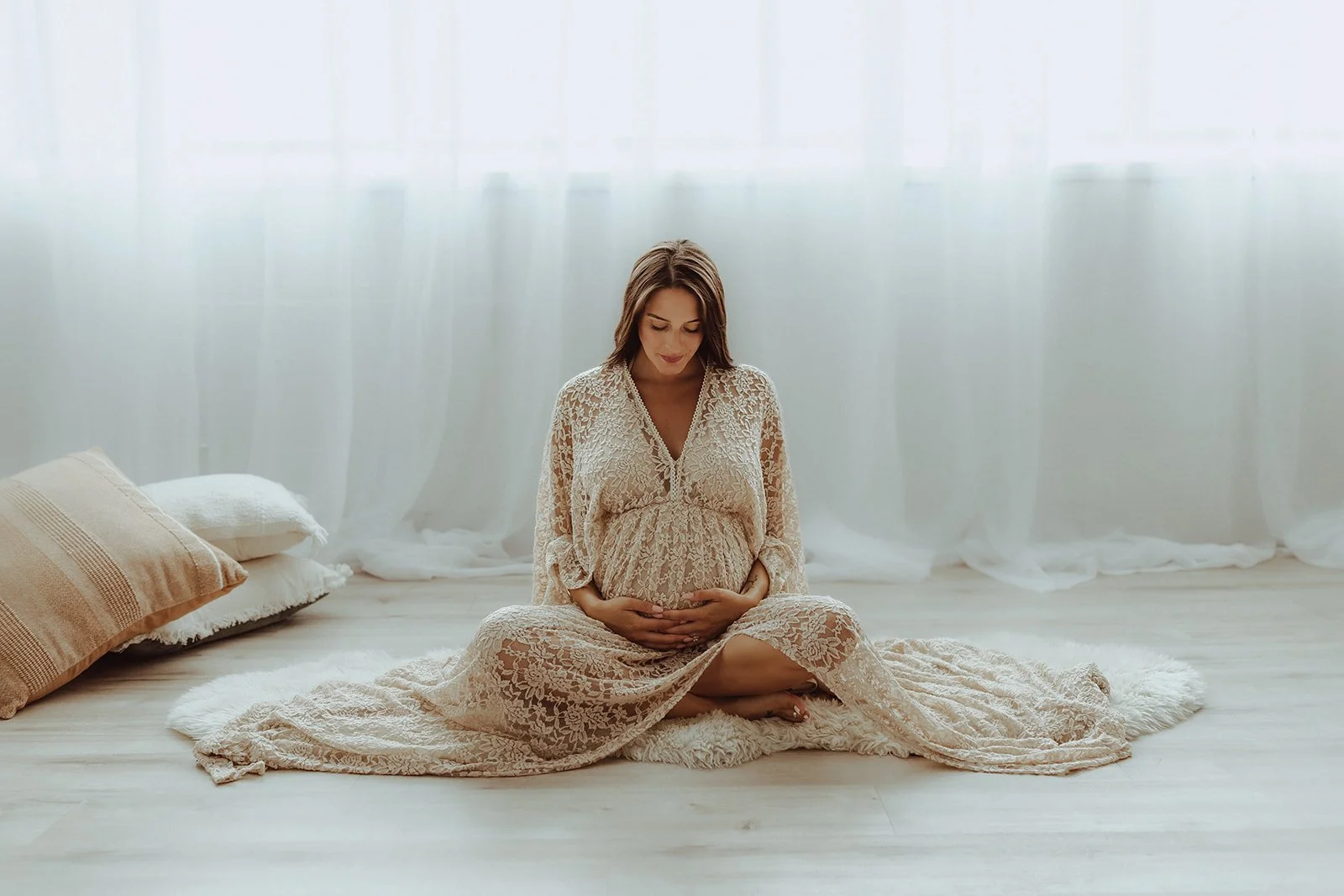 Pregnant woman sitting cross-legged on a soft rug, gently cradling her baby bump, in a bright room with sheer curtains.