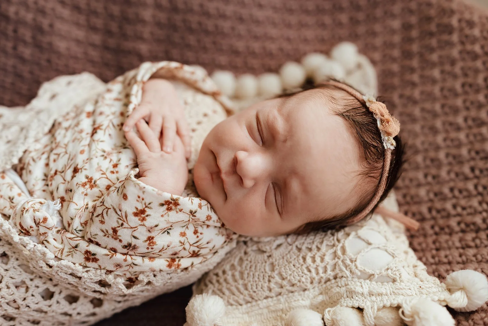 A sleeping baby with a floral headband, wrapped in a patterned blanket, lying on a textured cream-colored blanket on a brown surface.