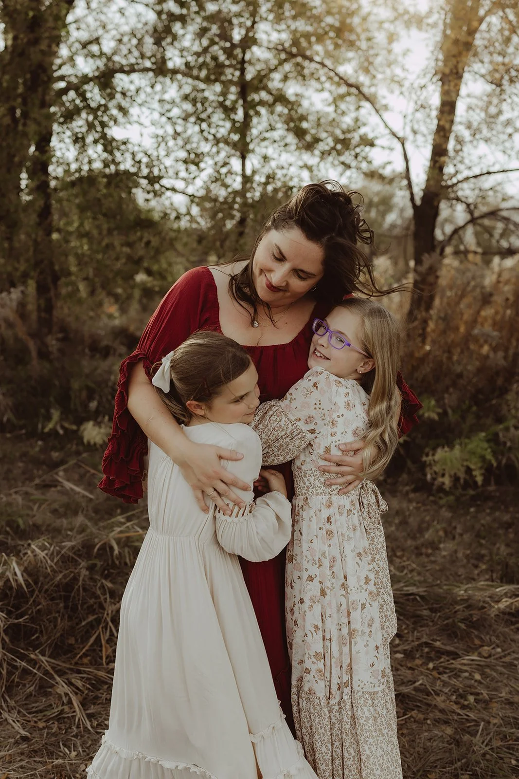 A woman hugging two young girls outdoors with trees and autumn foliage in the background.