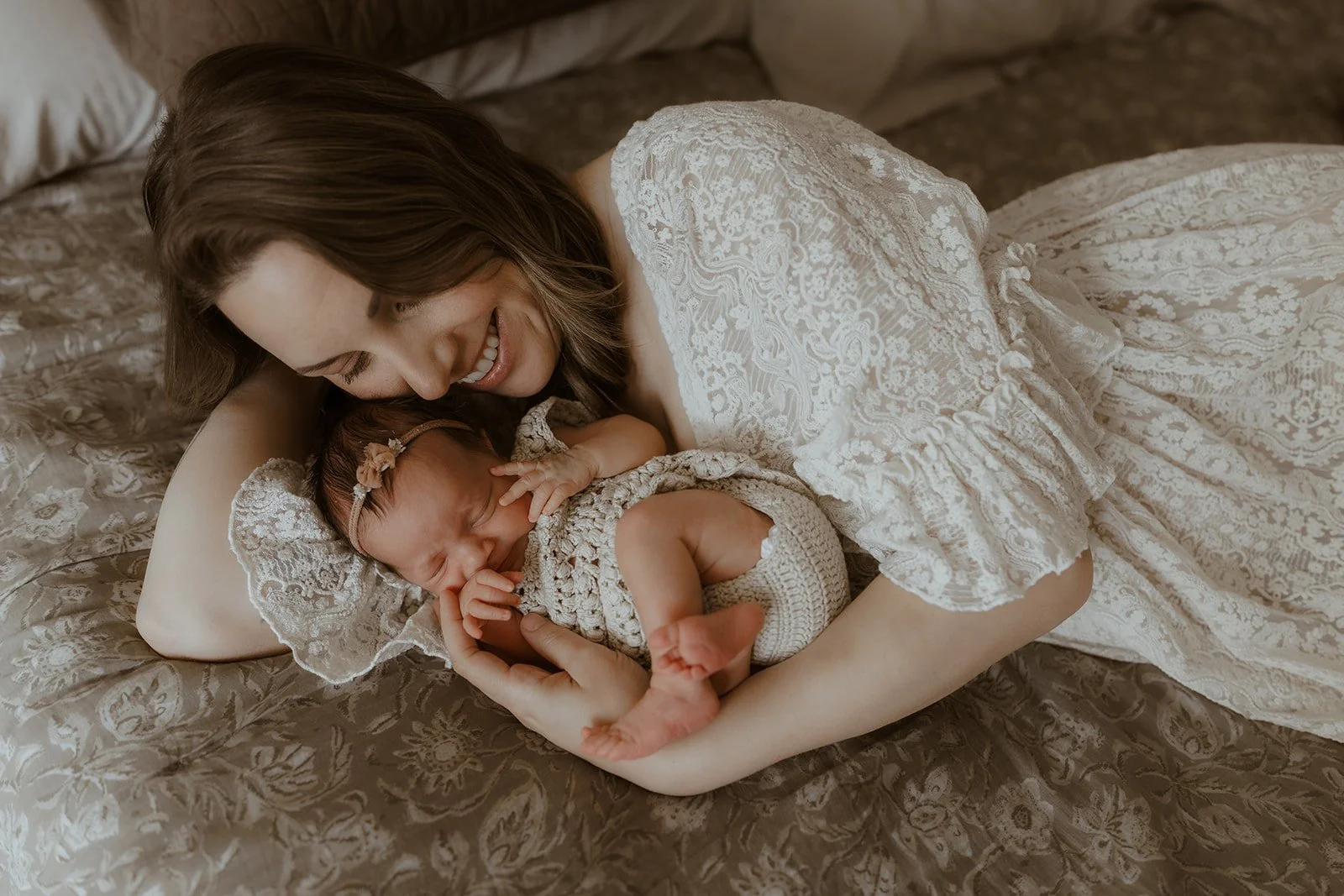 A woman with brown hair lying on a bed, smiling, embracing a sleeping baby girl dressed in a crochet outfit, on a bed with gold and beige patterned bedding.