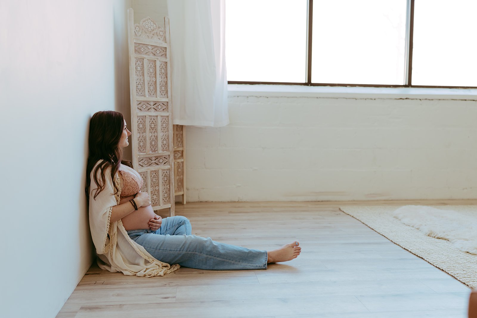 a woman wearing a bralette, jeans, and a cardigan is sitting on the floor during her maternity photo shoot with Ann Marie from AMG Photography
